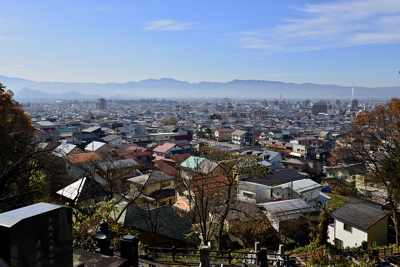 The rooftops of Aizuwakamatsu spread out below Iimoriyama, seen past gravestones on the hillside, with distant ranges on the horizon