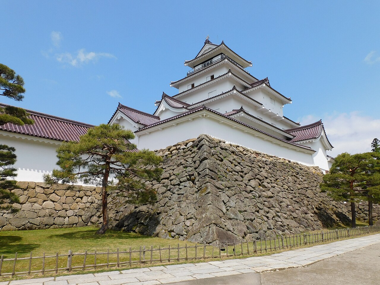 The white keep of Tsuruga Castle in Aizuwakamatsu rising above its dry-laid stone base, with pines and a gravel path in front under a clear sky