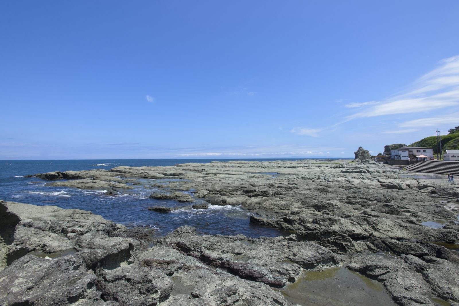 A flat rocky shoreline of dark rock platforms meeting the blue sea, with a few buildings in the distance under a clear sky.