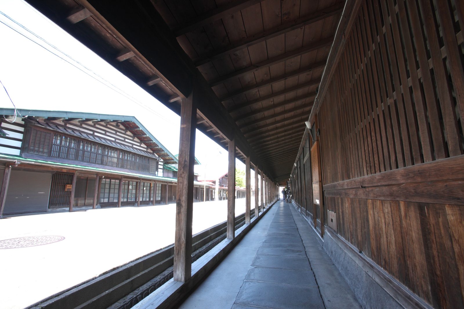 Looking down the covered wooden Komise arcade on Nakamachi Street in Kuroishi, with a stone walkway, a row of timber posts and a single figure far down the street