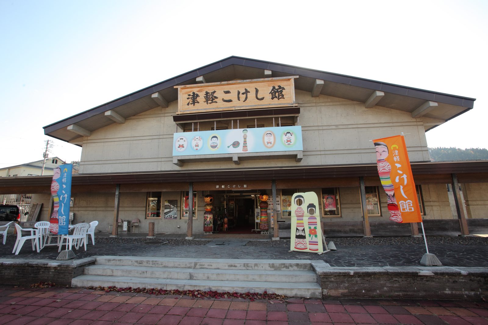 Exterior of the Tsugaru Kokeshi Museum in Kuroishi, a low concrete and timber building with a carved wooden sign and tall kokeshi banners beside the entrance
