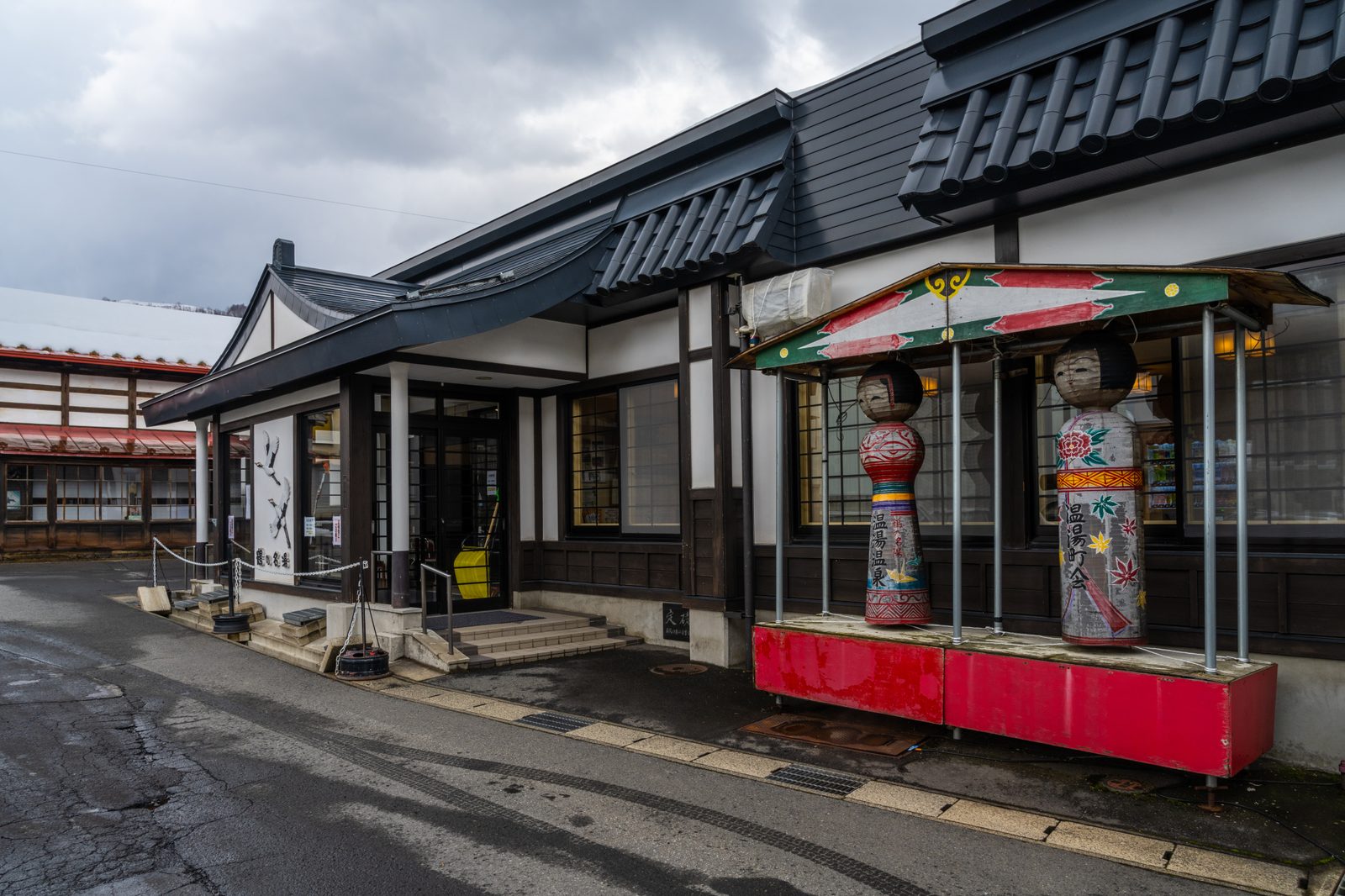 Entrance of the public bathhouse at Nuruyu Onsen in Kuroishi, with two tall painted kokeshi figures standing under a small canopy beside it on an overcast winter day