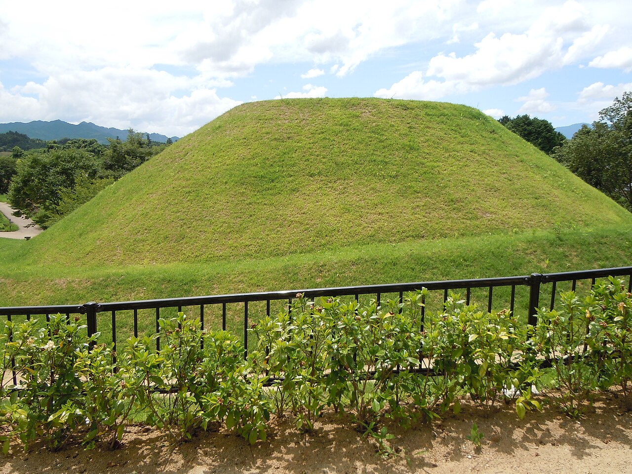The grass-covered round mound of the Takamatsuzuka tomb in Asuka, seen over a low railing with wooded hills behind it.