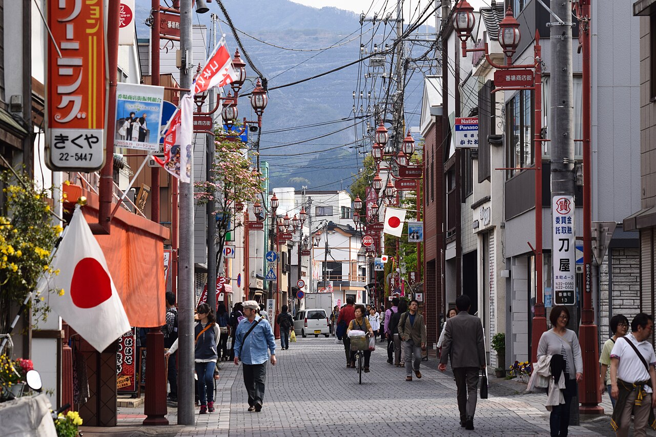 Low shops, red street lamps and hanging banners line the paved Banba shopping street in central Chichibu, with a wooded ridge at the far end.