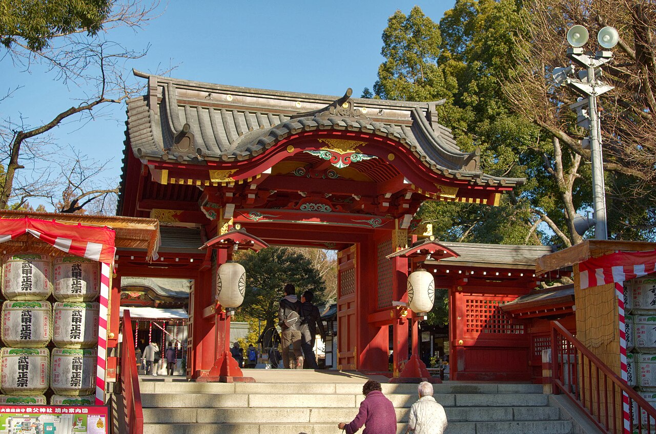 The vermilion gate of Chichibu Shrine stands under a clear sky, with white paper lanterns hung in the gateway and stacked sake barrels to either side.
