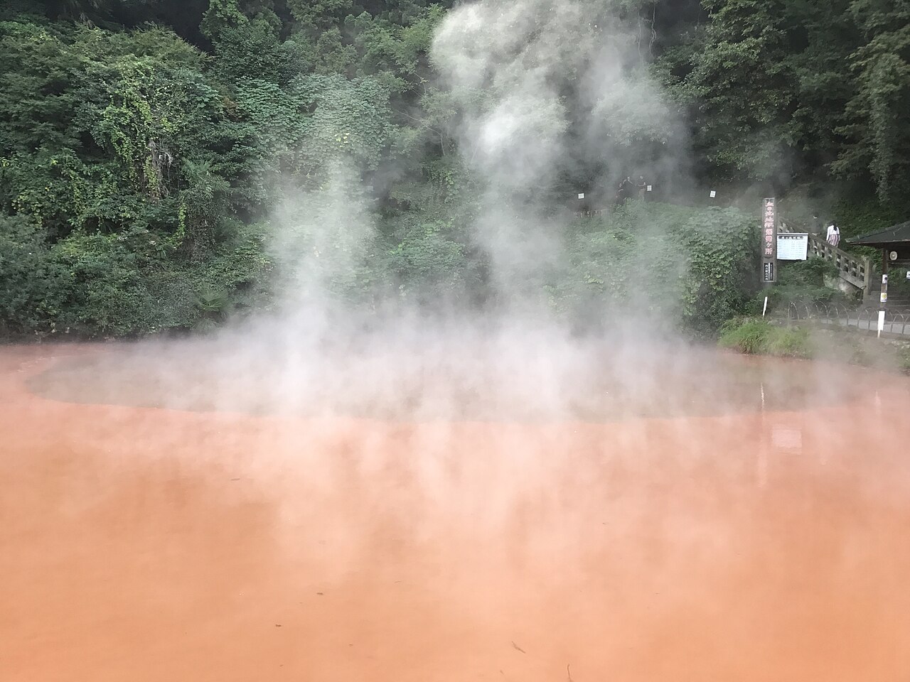 Steam drifting across the red-ochre water of Chinoike Jigoku in Beppu, ringed by dense green hillside and a wooden viewing rail.