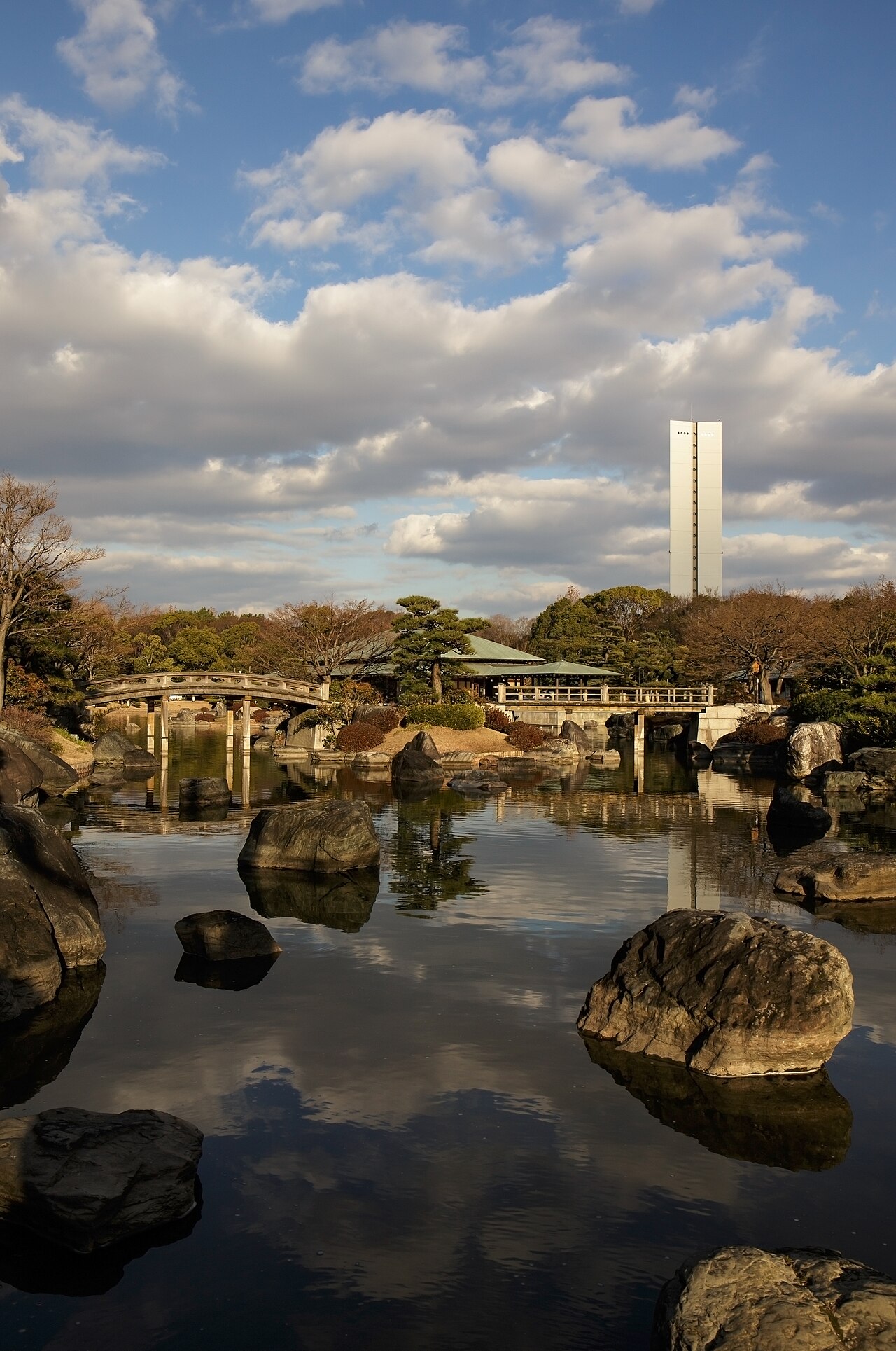 A quiet pond in the Daisen Park Japanese garden, with a low arched bridge, a green-roofed pavilion, and a slender white tower above the trees