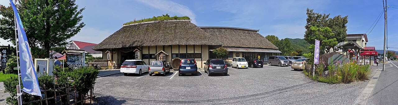 The thatched-roof main hall of Denshoen folk village in Tono, with parked cars and a blue banner flag out front on a sunny day.