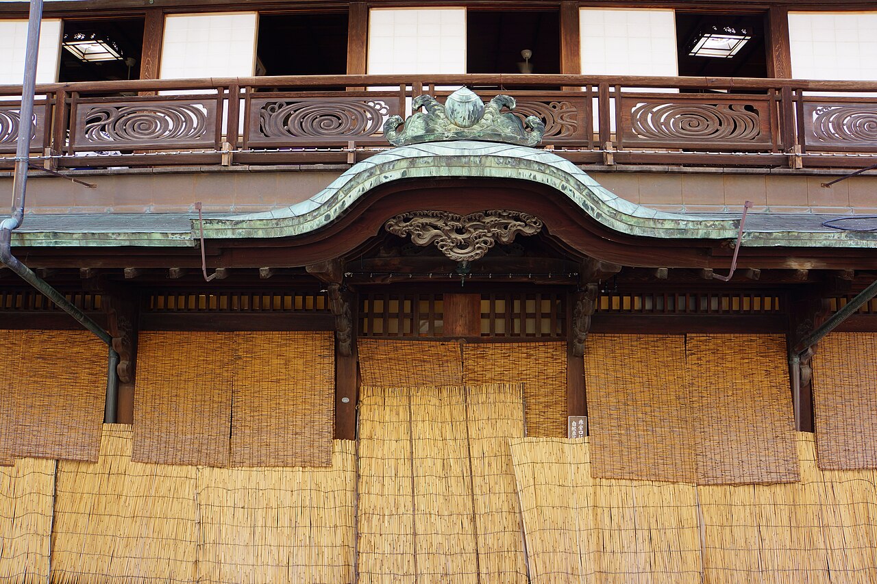 A close-up of the ornate curved gable and copper roof above the entrance of Dogo Onsen Honkan, with carved wooden railings and reed screens below.