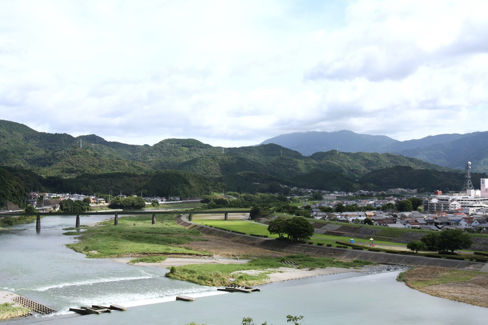 A wide view down over a river with a long low bridge, a grassy riverside park with a few trees, town buildings on the right and forested mountains under a cloudy sky.