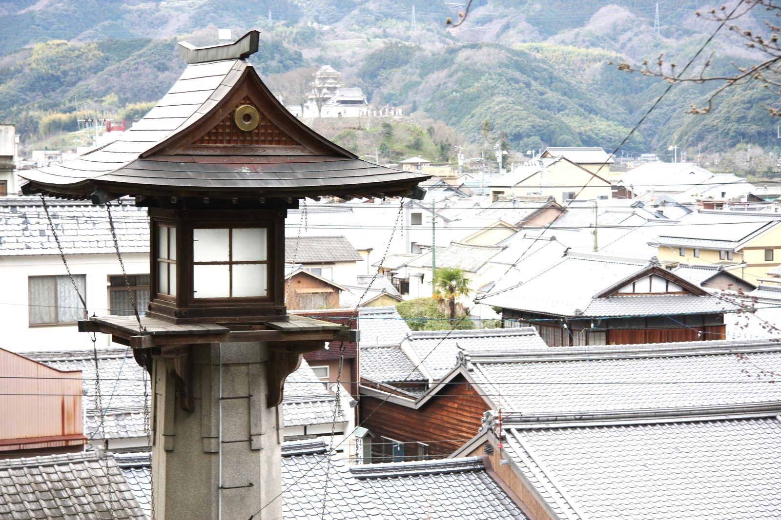 A wooden lantern with a curved tiled roof stands above the gray tiled rooftops of an old town, with a small castle keep on a hill and forested mountains behind.