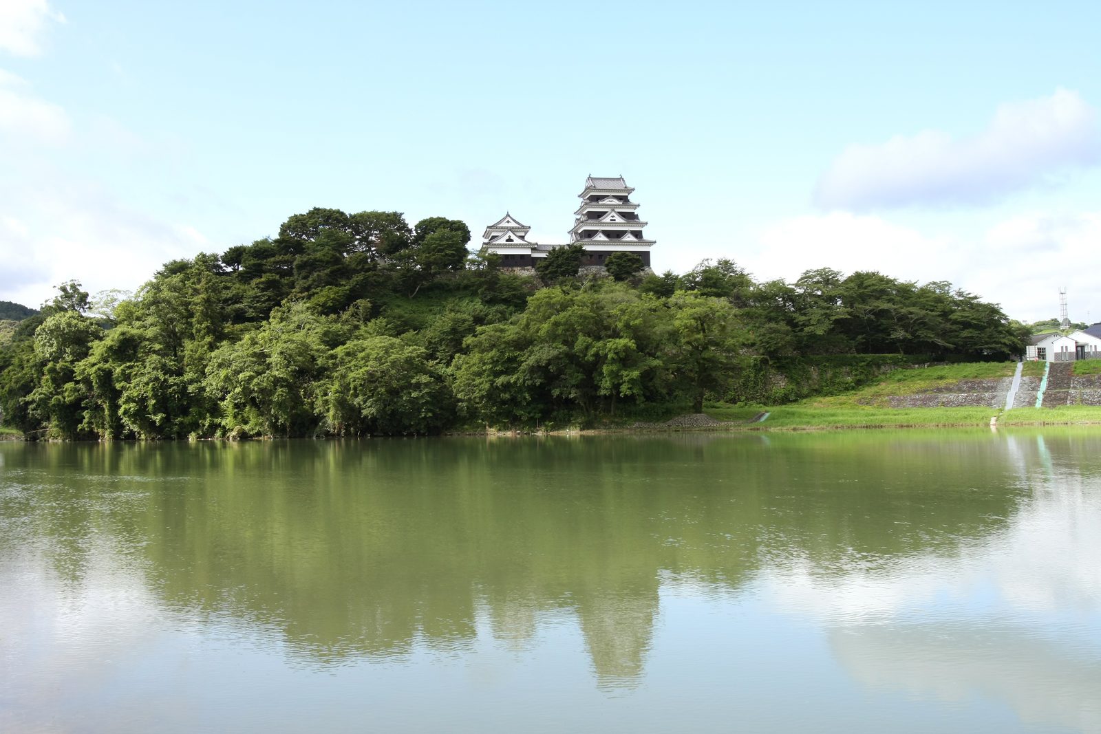 Ozu Castle's white keep and a smaller turret on a tree-covered hill above a wide, still river, with the hill reflected in the water under a blue sky.
