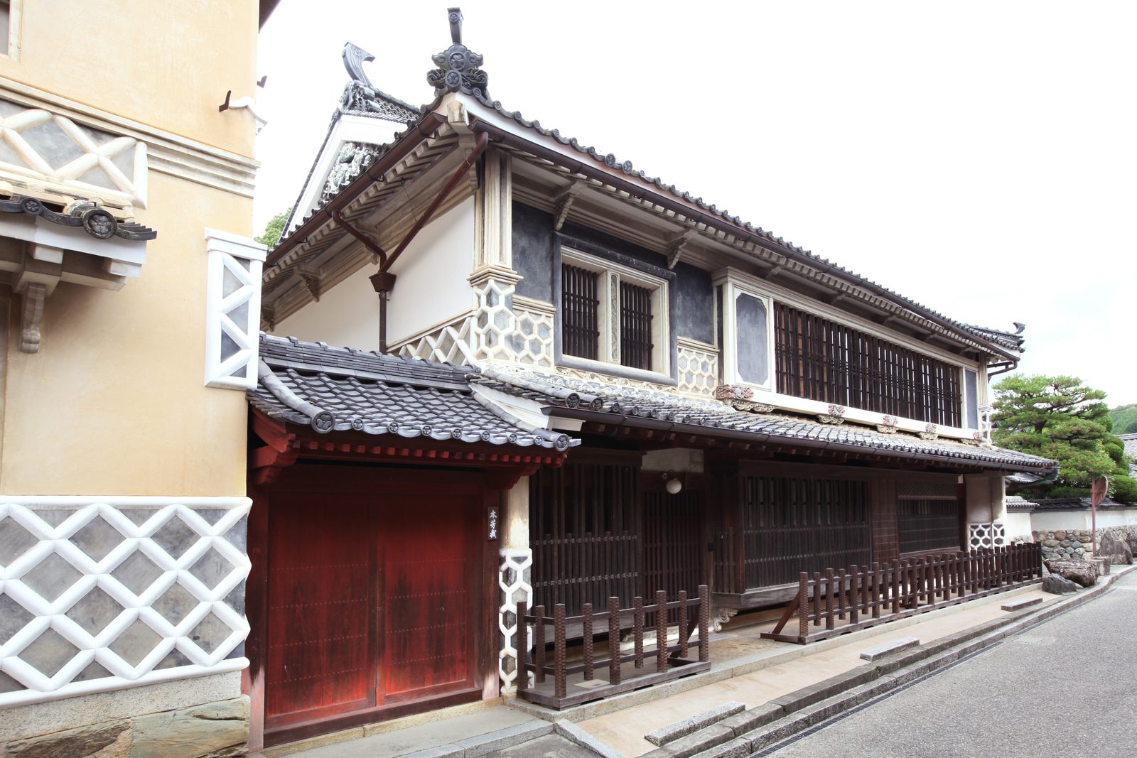 A two-storey merchant house with lattice windows, elaborate white plaster ornament and a red wooden gate, beside a paved street