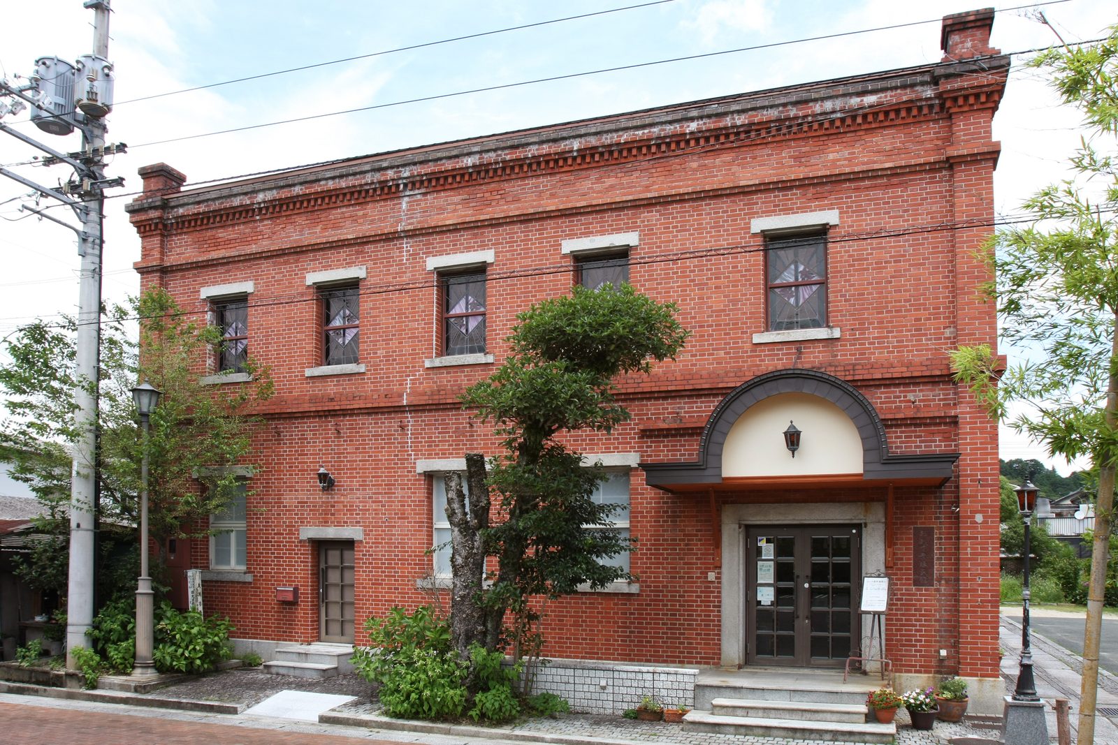 The red-brick facade of a two-story building with granite-framed windows, an arched entrance porch and a lamp post, on a paved street lined with small trees.