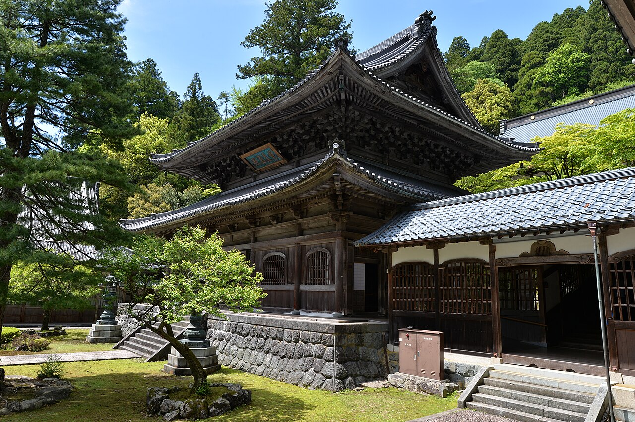 A dark timber Buddha hall with deeply bracketed eaves and a tiled roof stands on a stone plinth above a moss lawn, with cedar-covered hillside rising behind it at Eiheiji.
