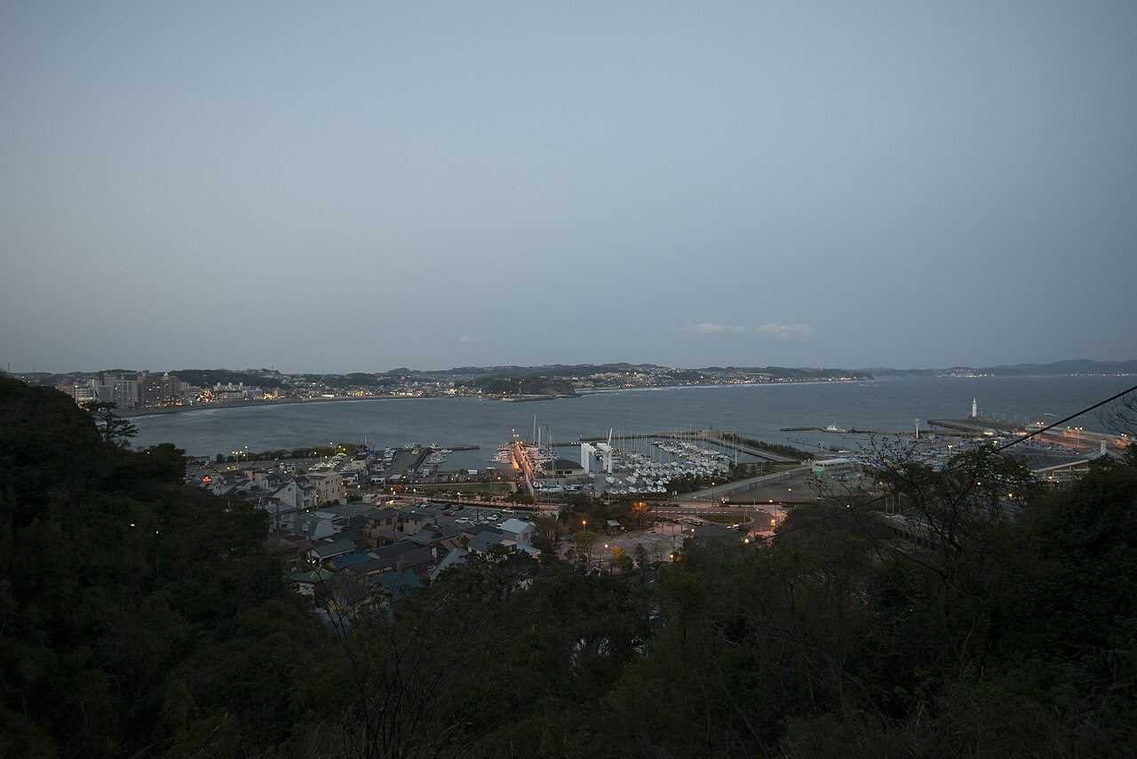 An elevated view over the small harbor and marina at the foot of Enoshima, with the curve of the Shonan coastline stretching into the distance.
