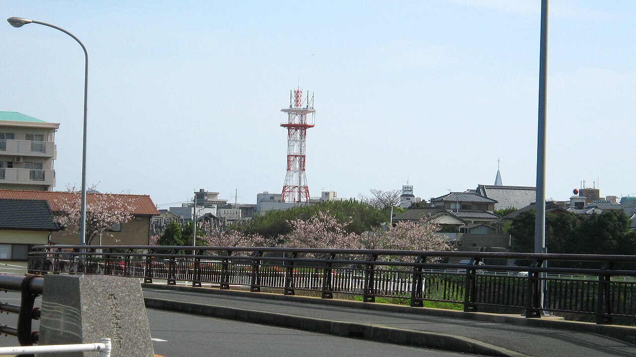 A road bridge in Fukue with cherry blossom along the far bank, tiled roofs and a red and white radio mast beyond