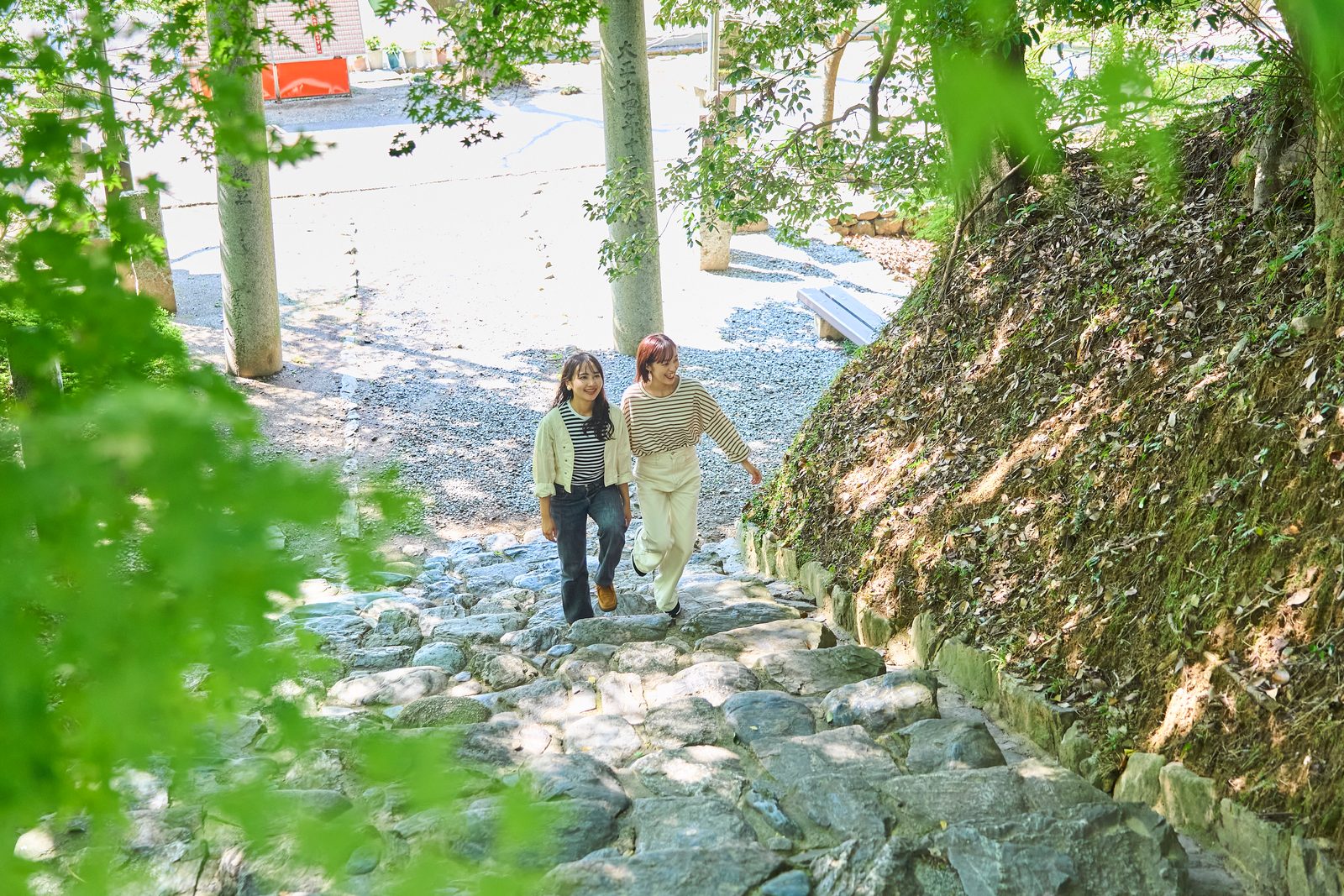 Two women in casual clothes walk down a mossy stone staircase toward a gravel courtyard with tall stone pillars and a wooden bench, framed by fresh green maple leaves overhead.