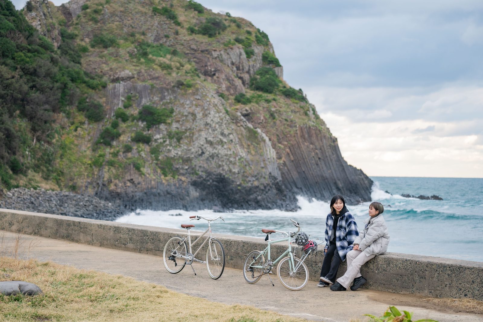 A rocky headland with columnar cliffs rising from the sea at Keya no Oto, with a seawall path and two parked bicycles in the foreground