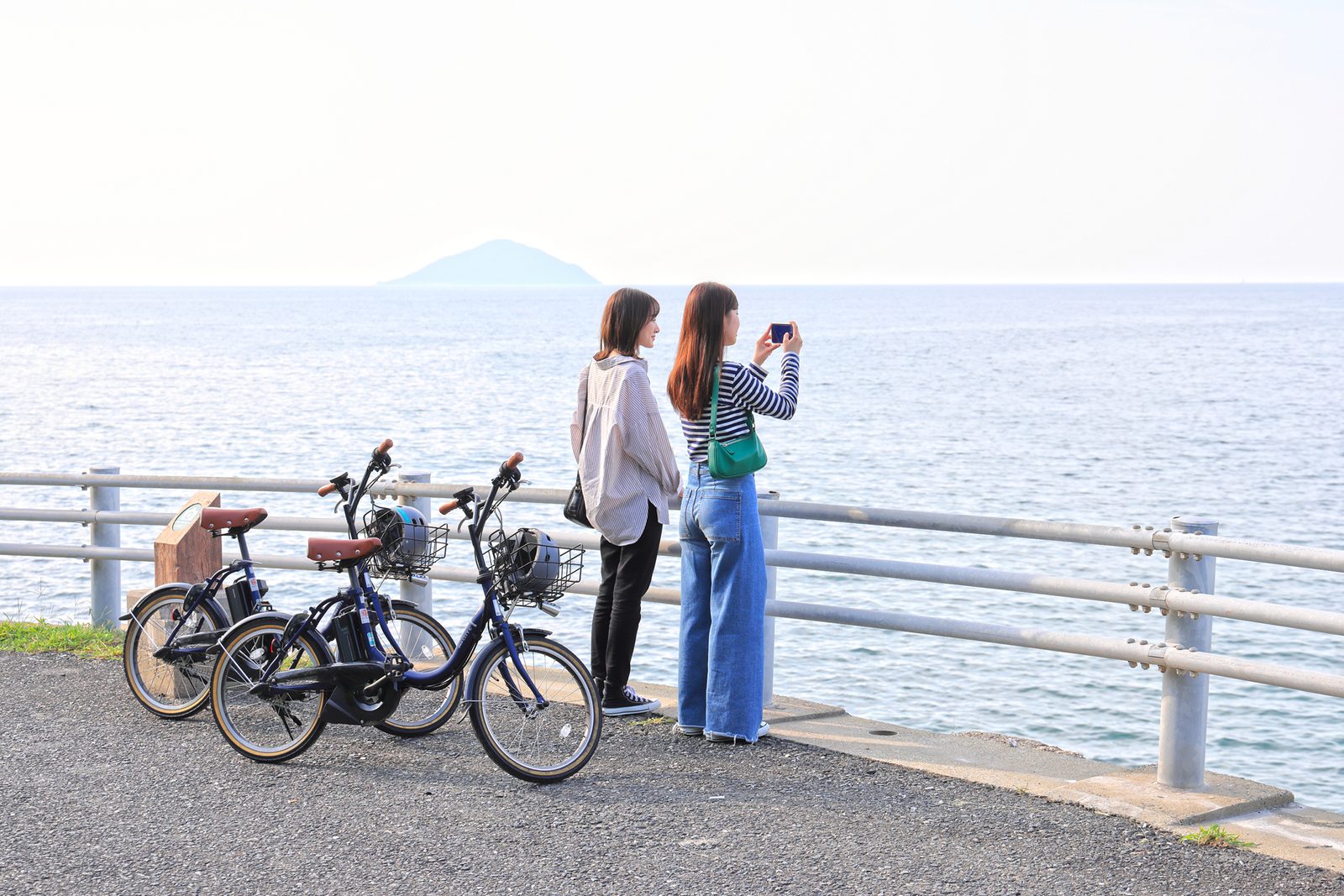 Two women with bicycles by a white guardrail photographing the calm sea, with a distant mountain on the horizon along the Itoshima coastline