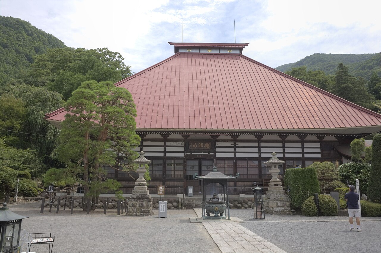 The broad tiled roof and wooden facade of the main hall at Ganshoin temple in Obuse, seen across its gravel forecourt with a bronze incense burner in front