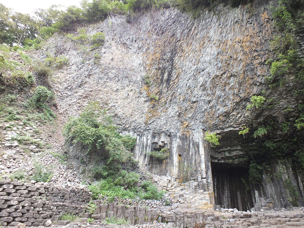 Tall gray basalt columns rise in tightly packed vertical ridges around a dark cave opening, with moss and ivy clinging to the weathered rock face.