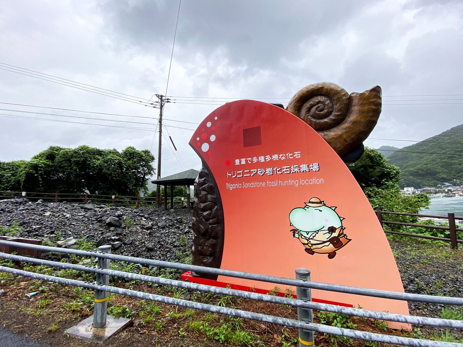 A red signboard topped with an ammonite sculpture marks the Trigonia sandstone fossil hunting location beside a gravel bed and the sea.
