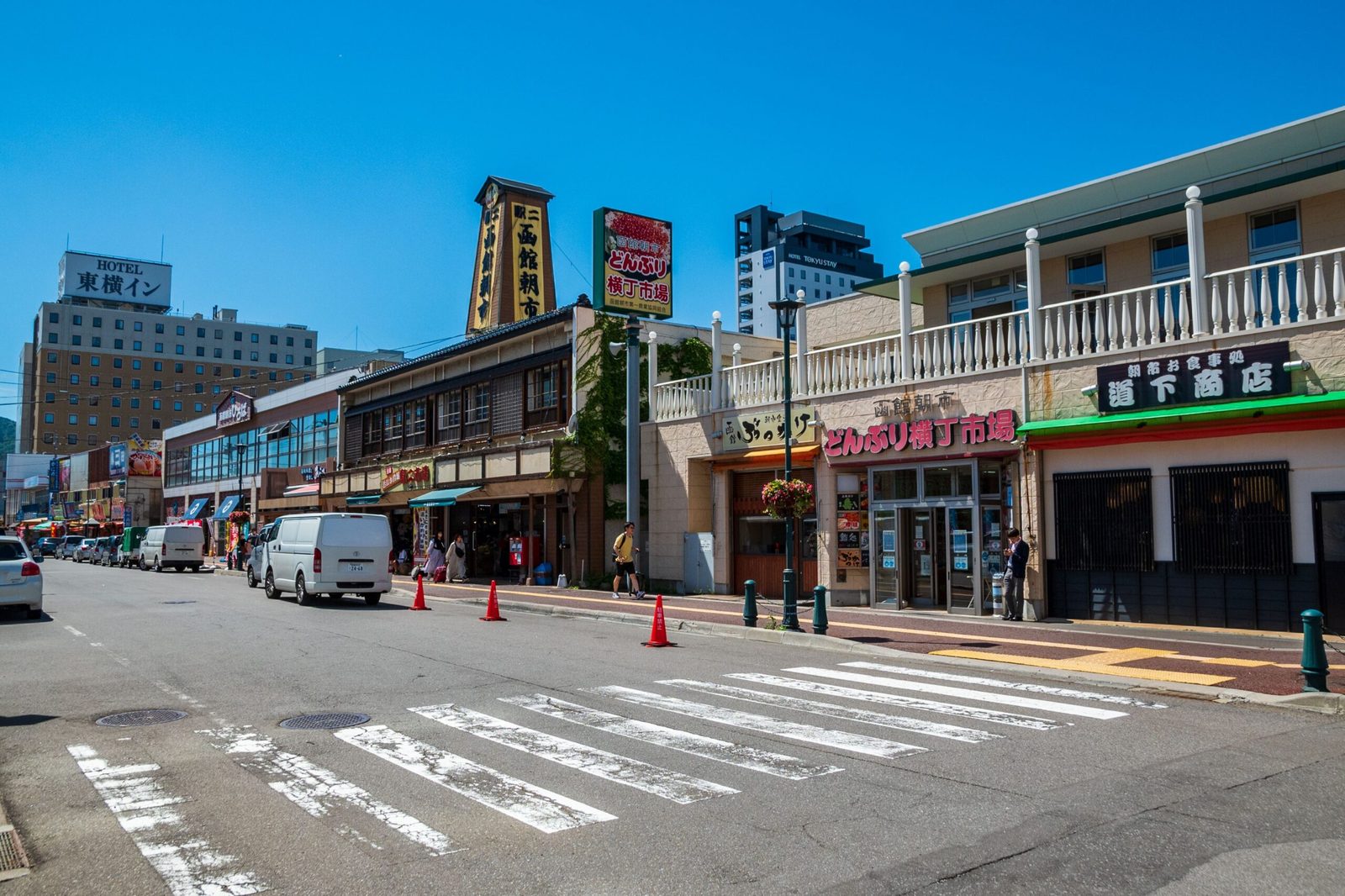 Street in front of the Hakodate Morning Market on a sunny day, with a tall wooden sign tower, the Donburi Yokocho market entrance, shops with signboards, a white van and orange traffic cones, and a crosswalk in the foreground