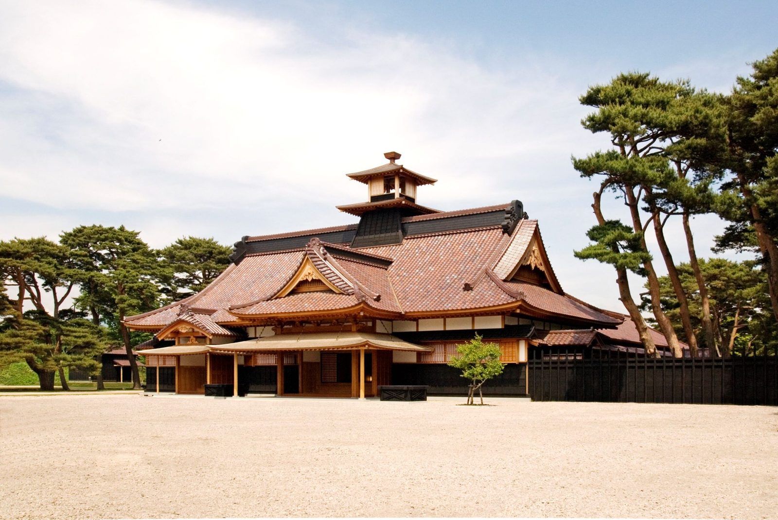 The reconstructed Hakodate Magistrate Office, a low building with a reddish-brown tiled roof and a small bell tower, seen across a wide gravel courtyard with pine trees and a black wooden fence under a partly cloudy sky