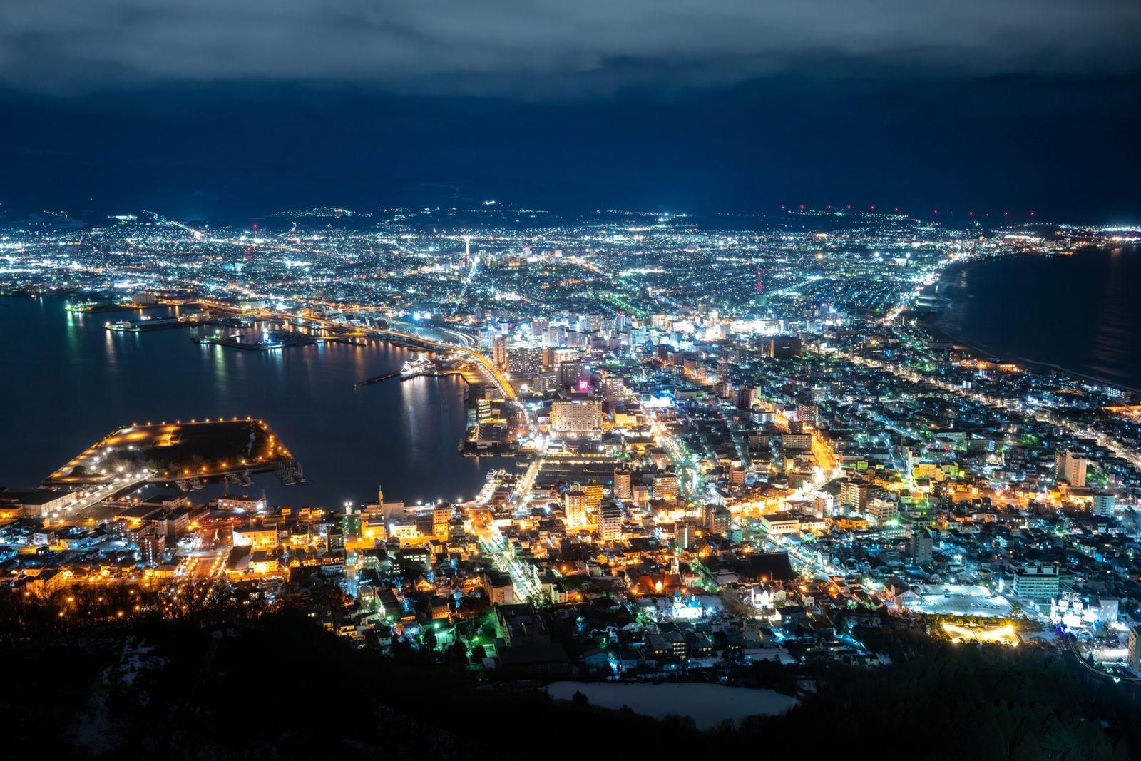 Night view of Hakodate from the summit of Mount Hakodate in winter, city lights spread across the narrow land between the bay and the sea, with patches of snow in the foreground and heavy cloud above