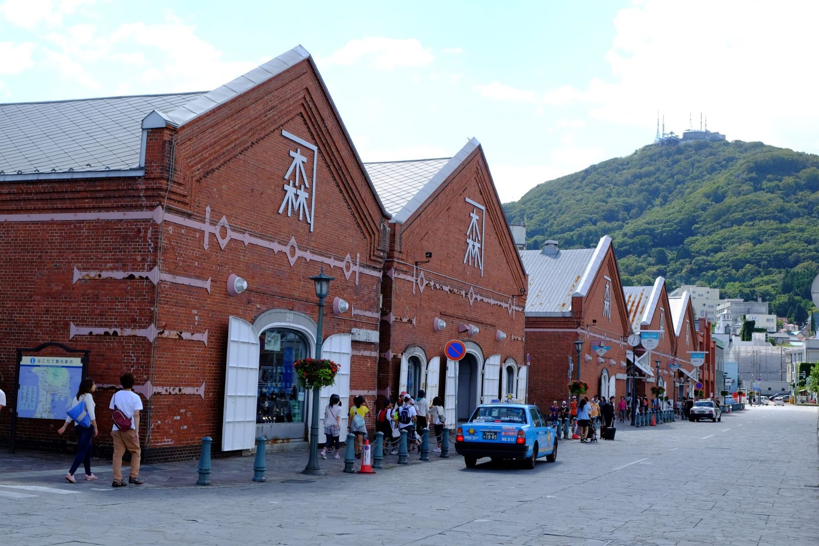 A row of red-brick warehouses with gabled fronts in Hakodate, people walking along the paved street, and the forested slope of Mount Hakodate with antenna towers on its summit behind
