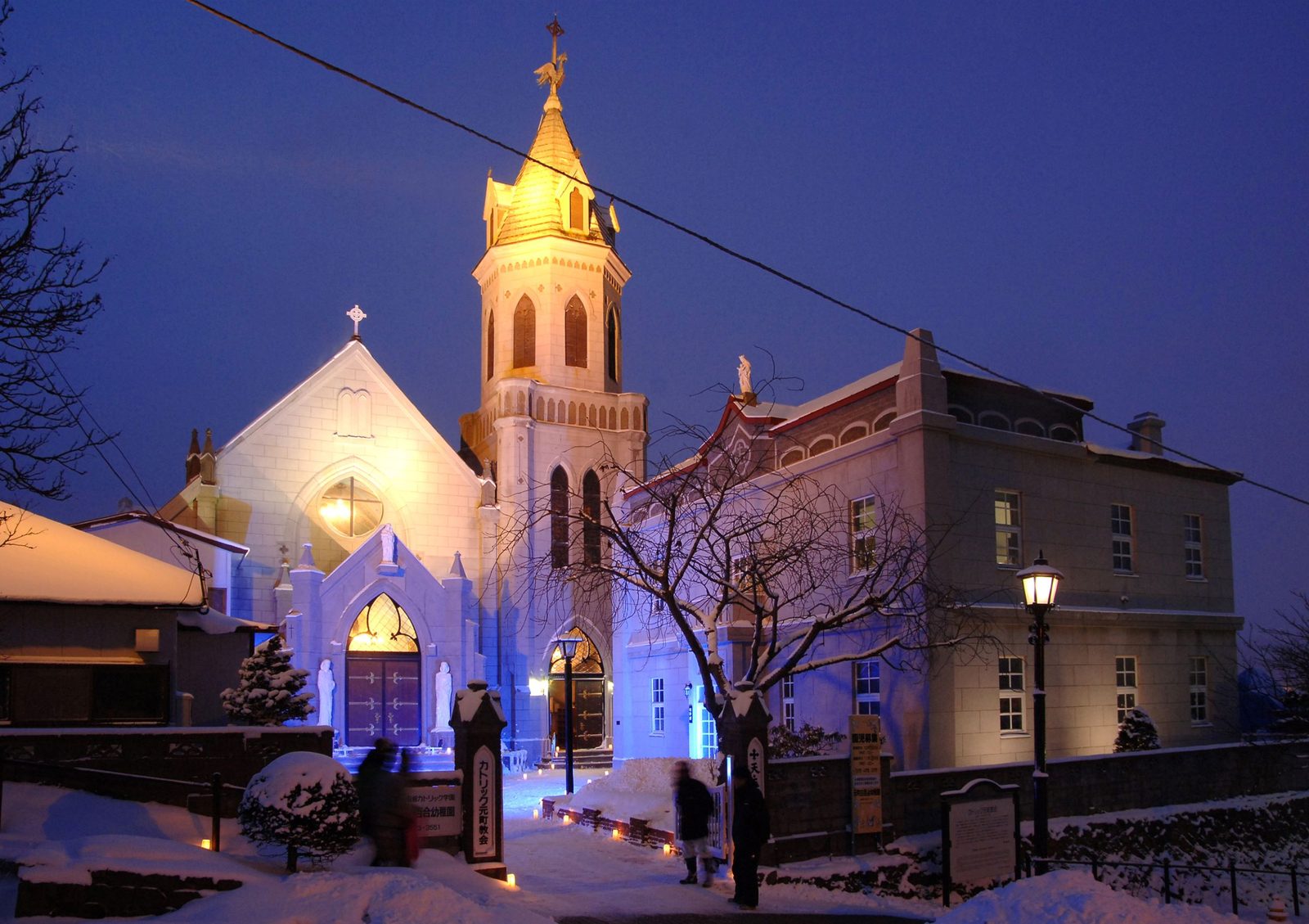 Catholic Motomachi Church in Hakodate at dusk in winter, its gold-lit bell tower and white gabled front glowing against a deep blue sky, with snow on the ground, small candle lights along the path, and a few people by the gate