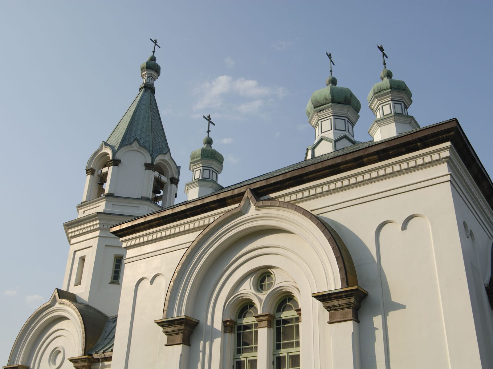 Close view of the Hakodate Orthodox Church, a white building with green copper onion-shaped domes topped with crosses, a bell tower on the left, and arched windows under a blue sky