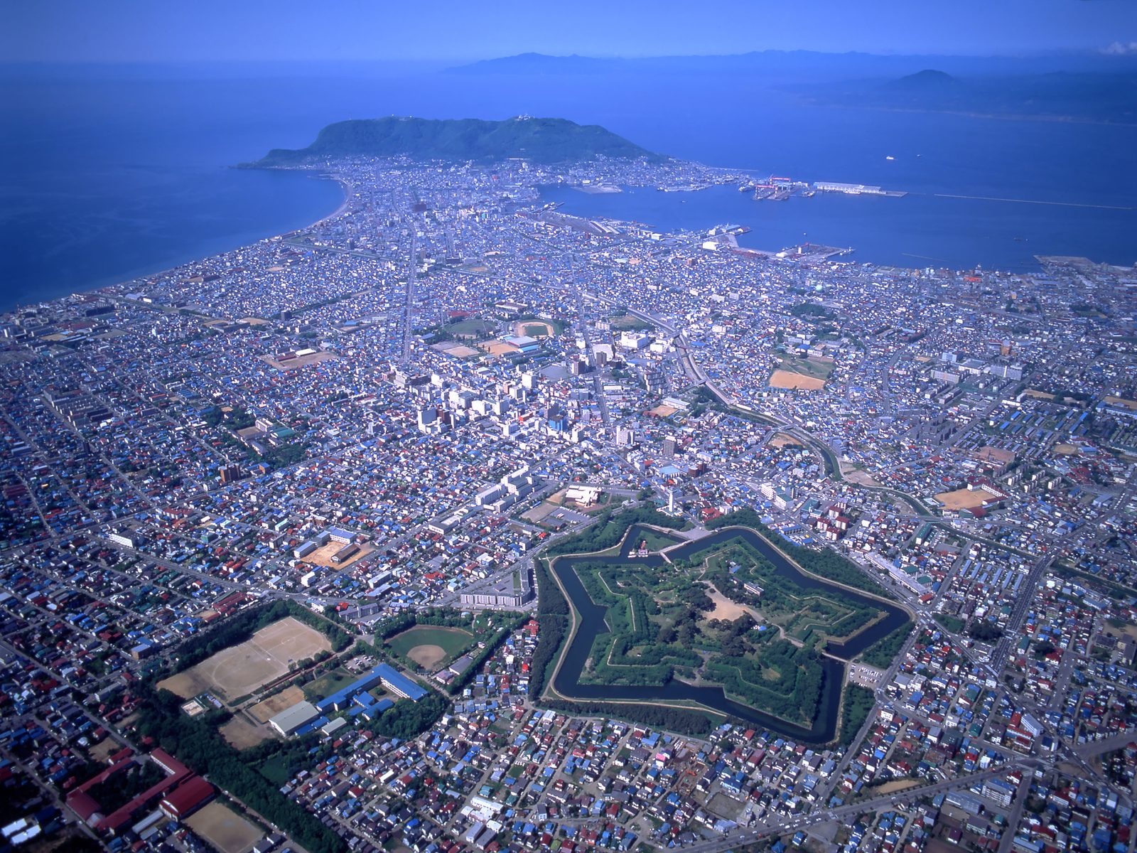 Aerial view of Hakodate in clear daylight, with the star-shaped Goryokaku fort and its moat in the foreground, the dense city in the middle, and the green bulk of Mount Hakodate at the end of the peninsula between the bays