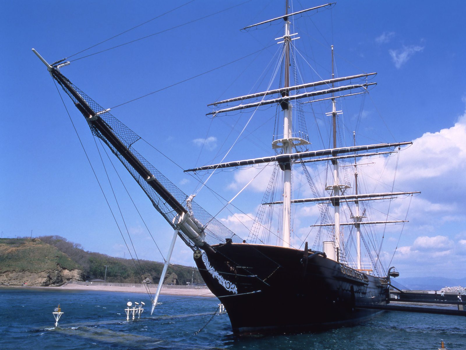 A reconstructed three-masted wooden sailing warship with a dark hull moored in a calm harbour under a bright blue sky.