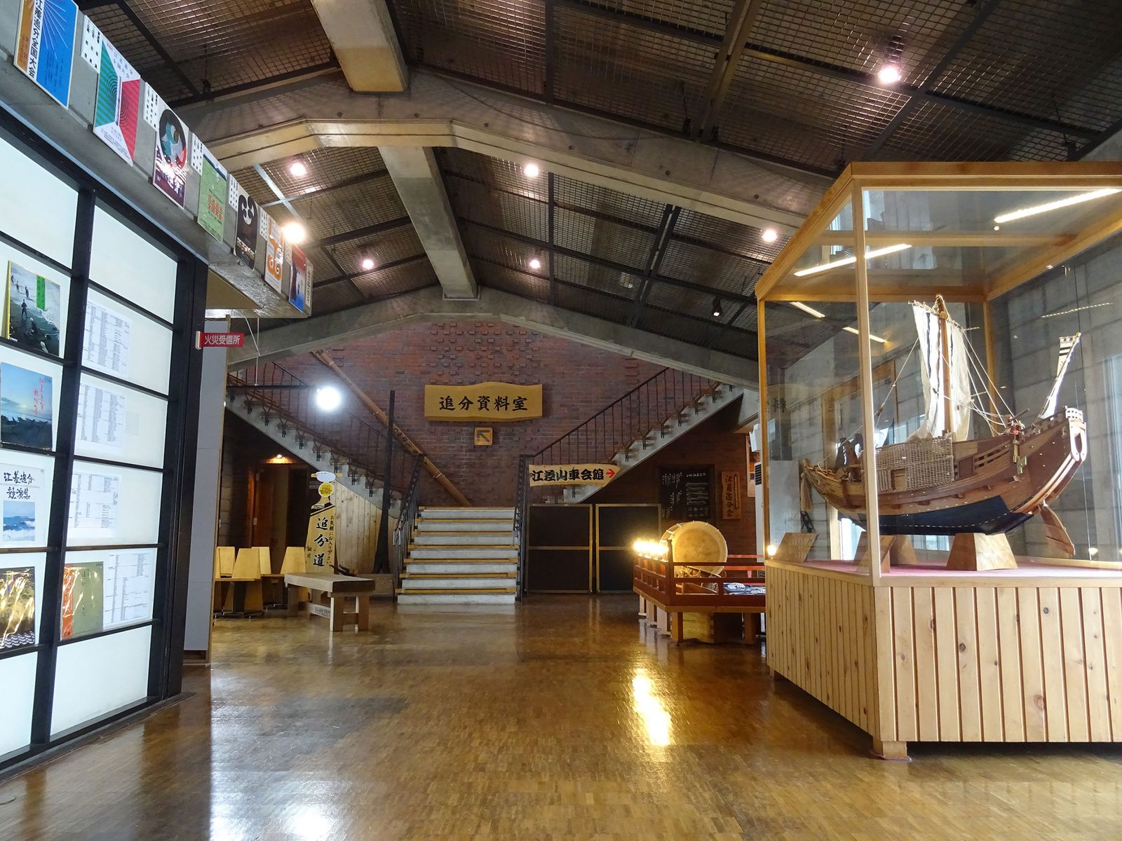 Inside a hall with a brick wall and a staircase, a glass case holds a wooden model of a sailing cargo ship beside a wall of posters and photographs.