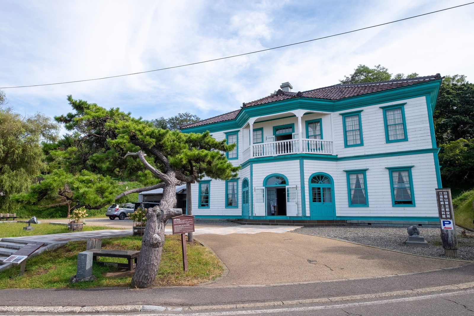 The white two-storey former Hiyama-Nishi district office building with turquoise trim and a balcony, now the Esashi Town local history museum, with a twisted pine tree in front.