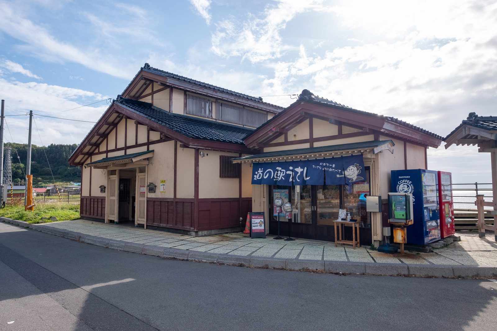 A traditional-style roadside station building with tiled roofs and a navy shop curtain at the entrance, with a vending machine and the sea beyond.