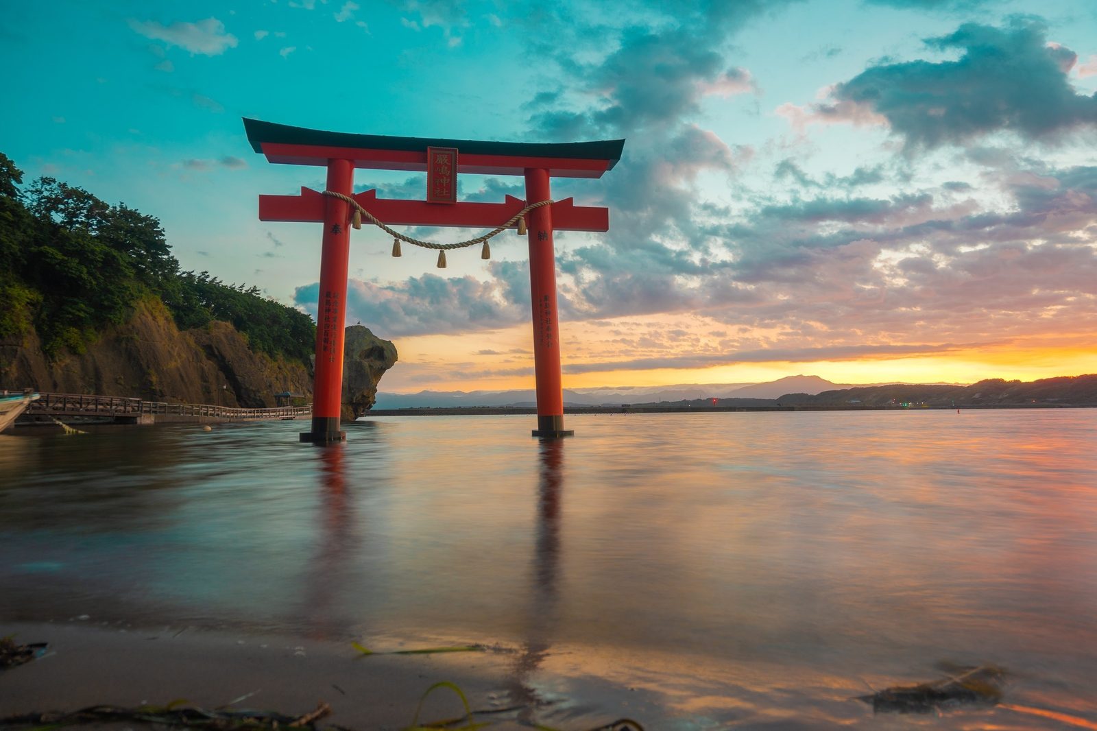 A red torii gate standing in shallow sea water at sunset, with colourful clouds, a rocky headland and distant hills.