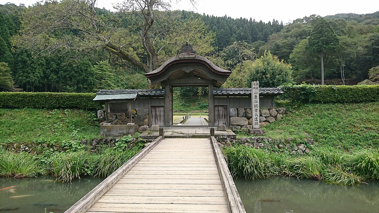 A small wooden gate with a curved cypress-bark roof sits in a clipped hedge and stone wall, reached by a plank footbridge over a narrow stream, with wooded hills behind it at the Ichijodani ruins.