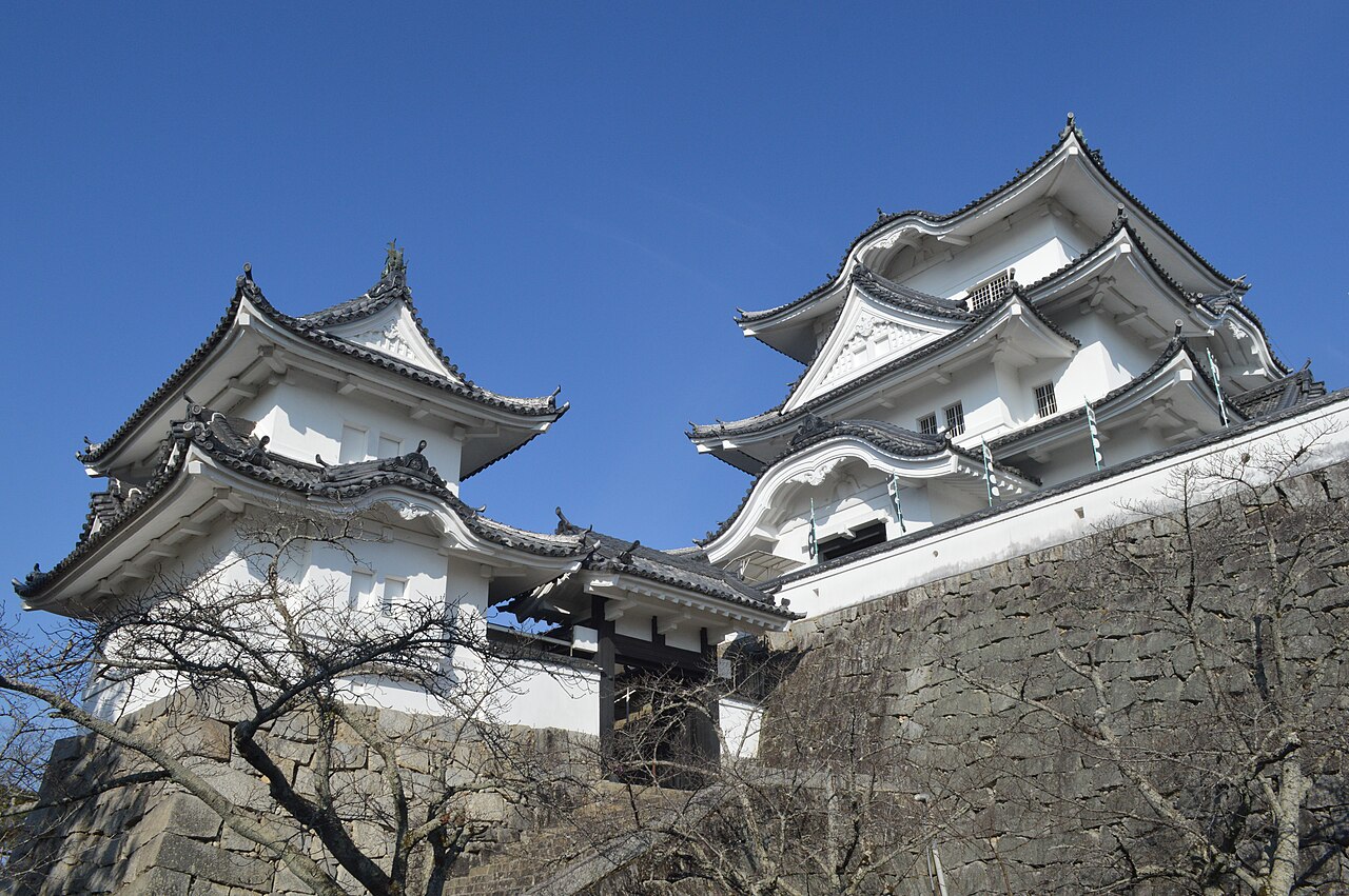 White main keep and small keep of Iga Ueno Castle seen from below, rising above a massive stone wall under a clear blue sky