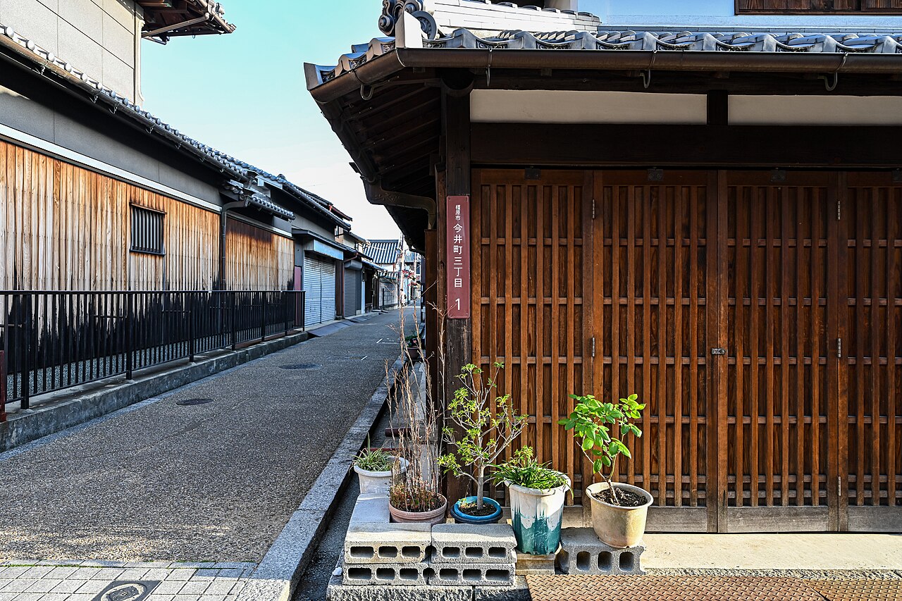 A narrow lane in Imai-cho lined with two-storey wooden merchant houses, white plaster upper walls, dark timber lattice fronts, and potted plants along the street.