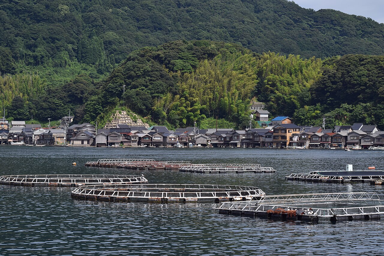 The village of Ine seen from across the bay, with fish pens in the foreground and wooded hills behind.