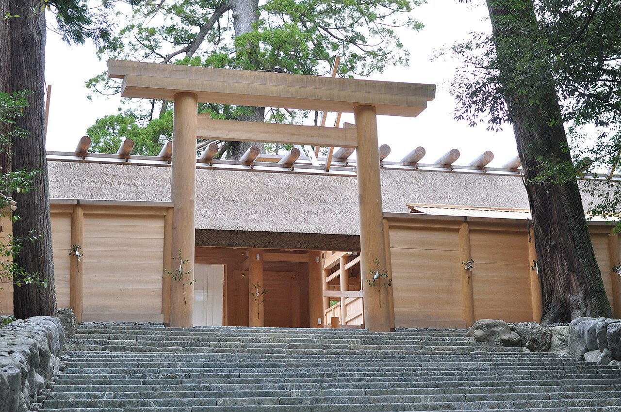 Broad stone steps rising to a plain wooden torii and the pale timber fence of the main sanctuary at the Inner Shrine of Ise, framed by cedars