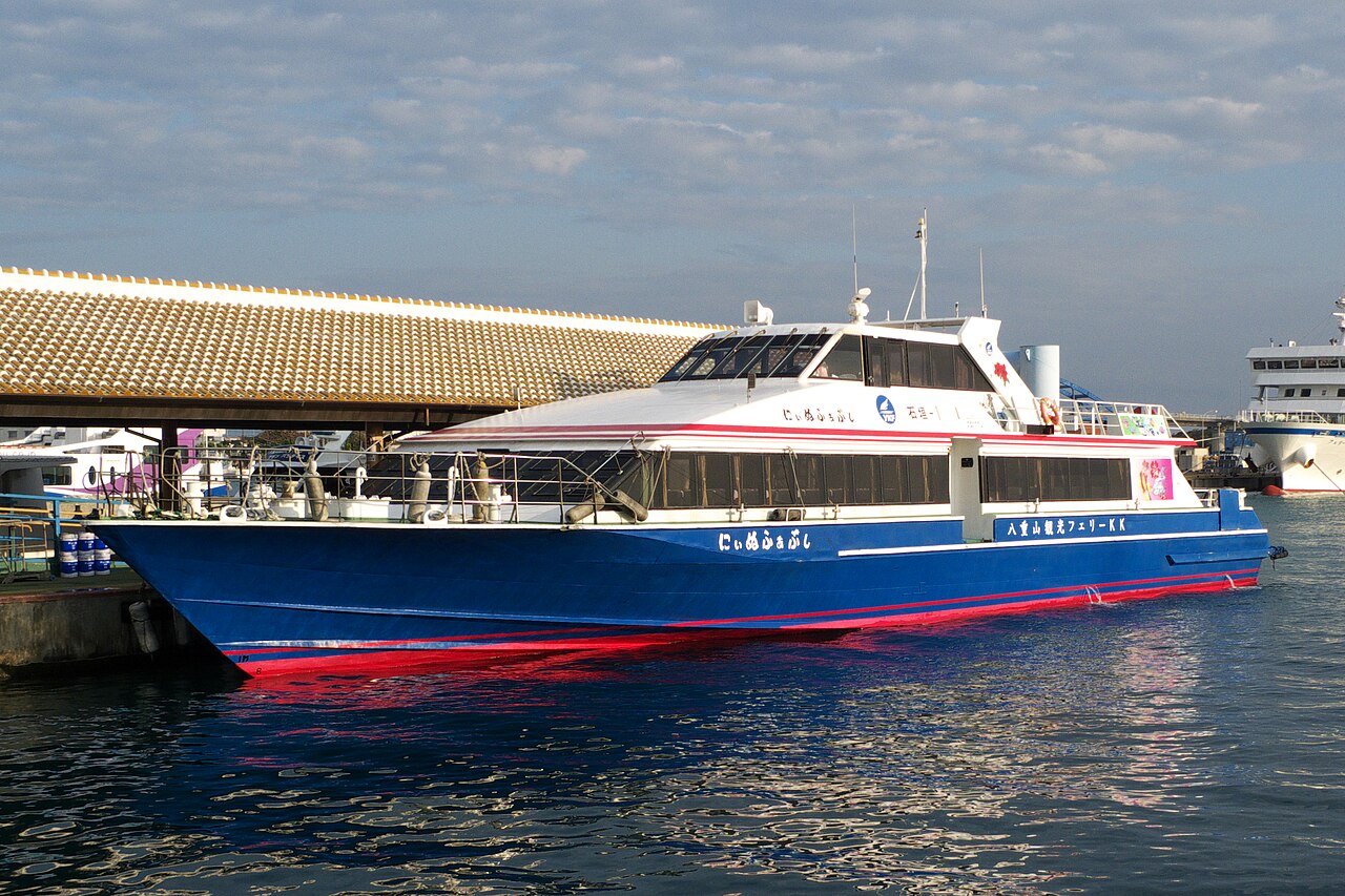 A blue and white high-speed boat moored at the Ishigaki Port terminal, with the terminal's tiled roof behind it