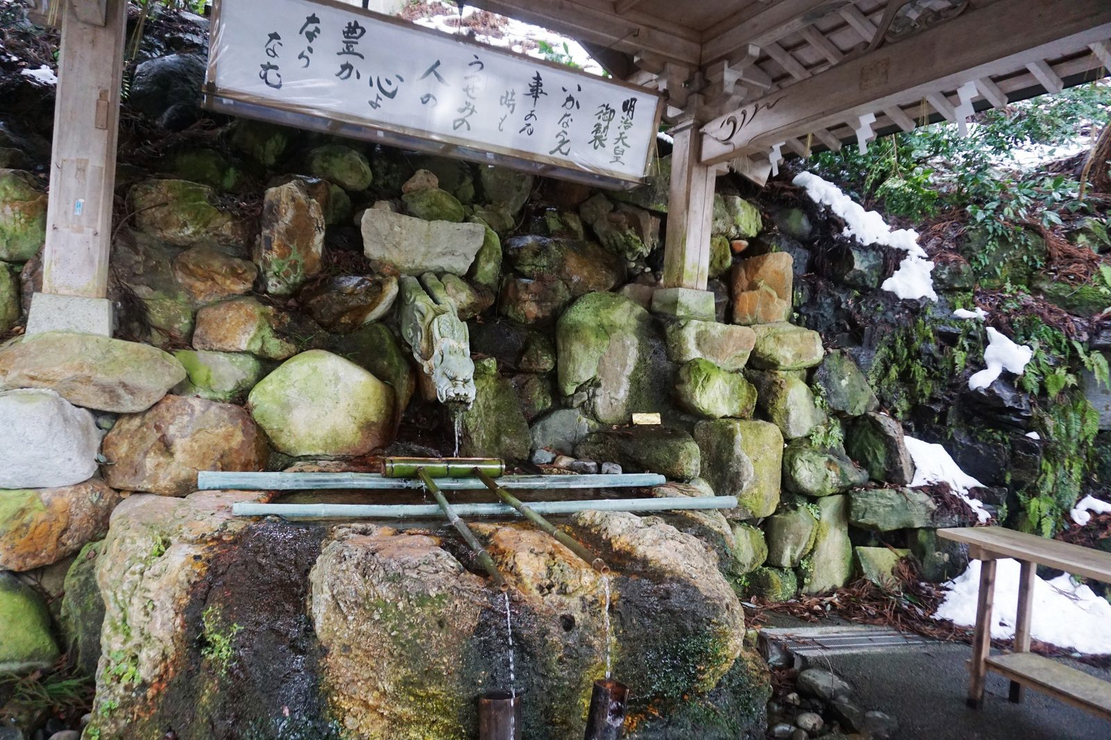 A stone temizuya purification fountain fed by a bronze dragon-head spout, with a bamboo ladle rest below and a wooden plaque of vertical Japanese text hanging above, set into a moss-covered rock wall.