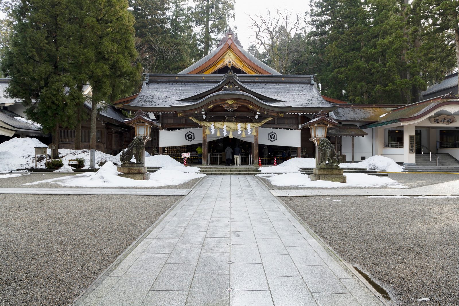 A wide view across the gravel courtyard toward the shrine's worship hall and an adjoining prayer hall building, framed by tall cedars with mounds of shoveled snow along the paved approach.