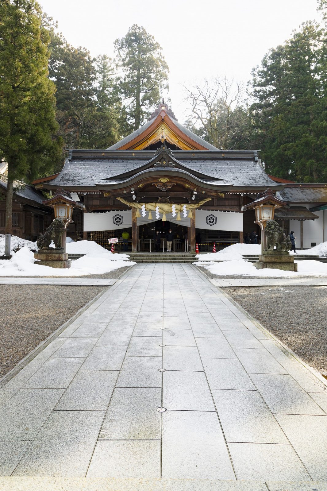The dark wood haiden worship hall of the shrine, its deep curved gable strung with a thick shimenawa rope and white paper streamers, flanked by bronze komainu guardian lions on stone pedestals with snowbanks piled at their base.