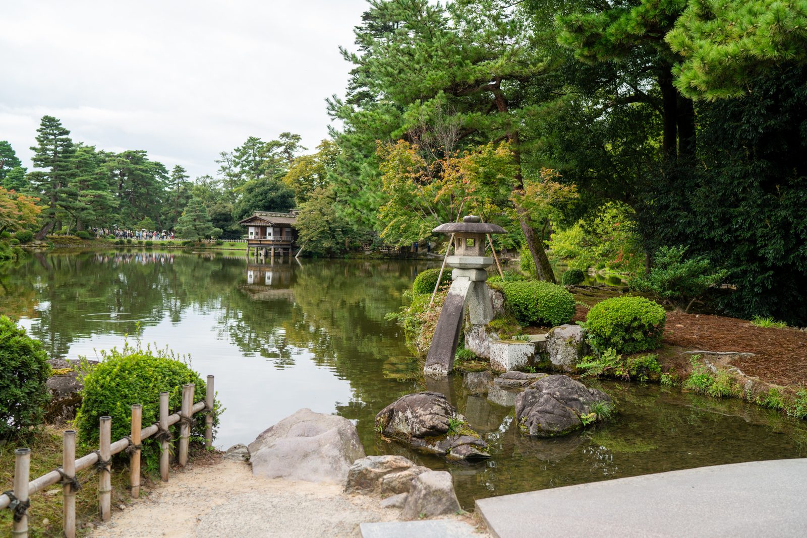 The two-legged stone Kotoji-toro lantern standing at the edge of a pond in Kenrokuen Garden, reflected in the still water with a wooden bridge and pine trees behind it.
