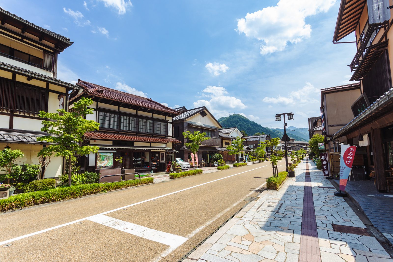 A quiet street lined with wooden-fronted shops and inns under a blue summer sky, with forested hills at the end of the road.